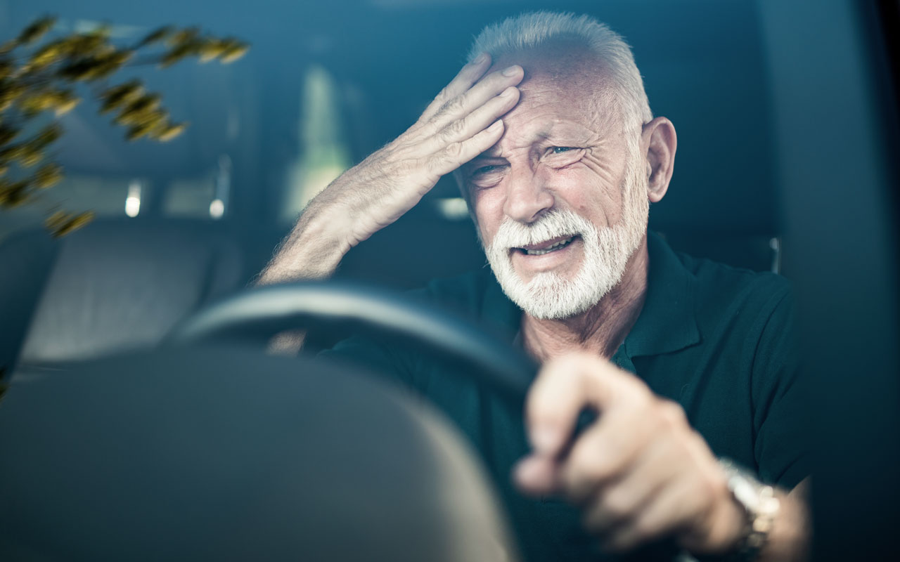 An elderly man behind the wheel of a vehicle appears distressed as he touches his forehead.